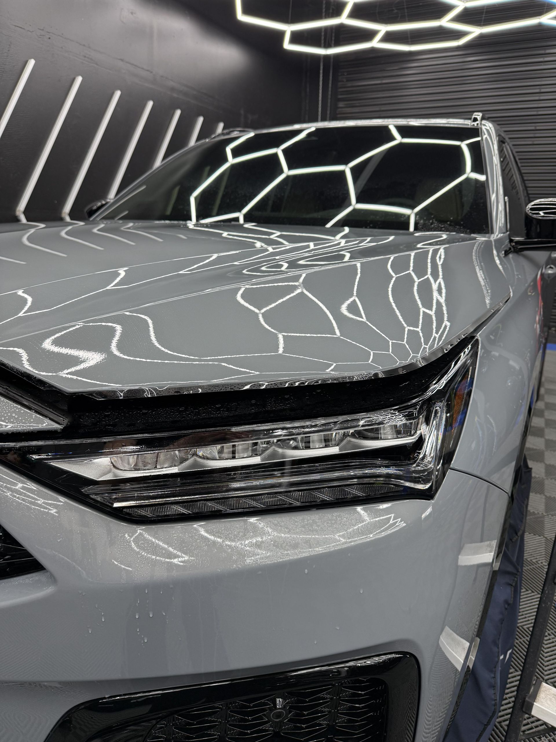 Grey SUV in a car detailing studio, reflecting overhead hexagonal lights, glistening with water droplets.