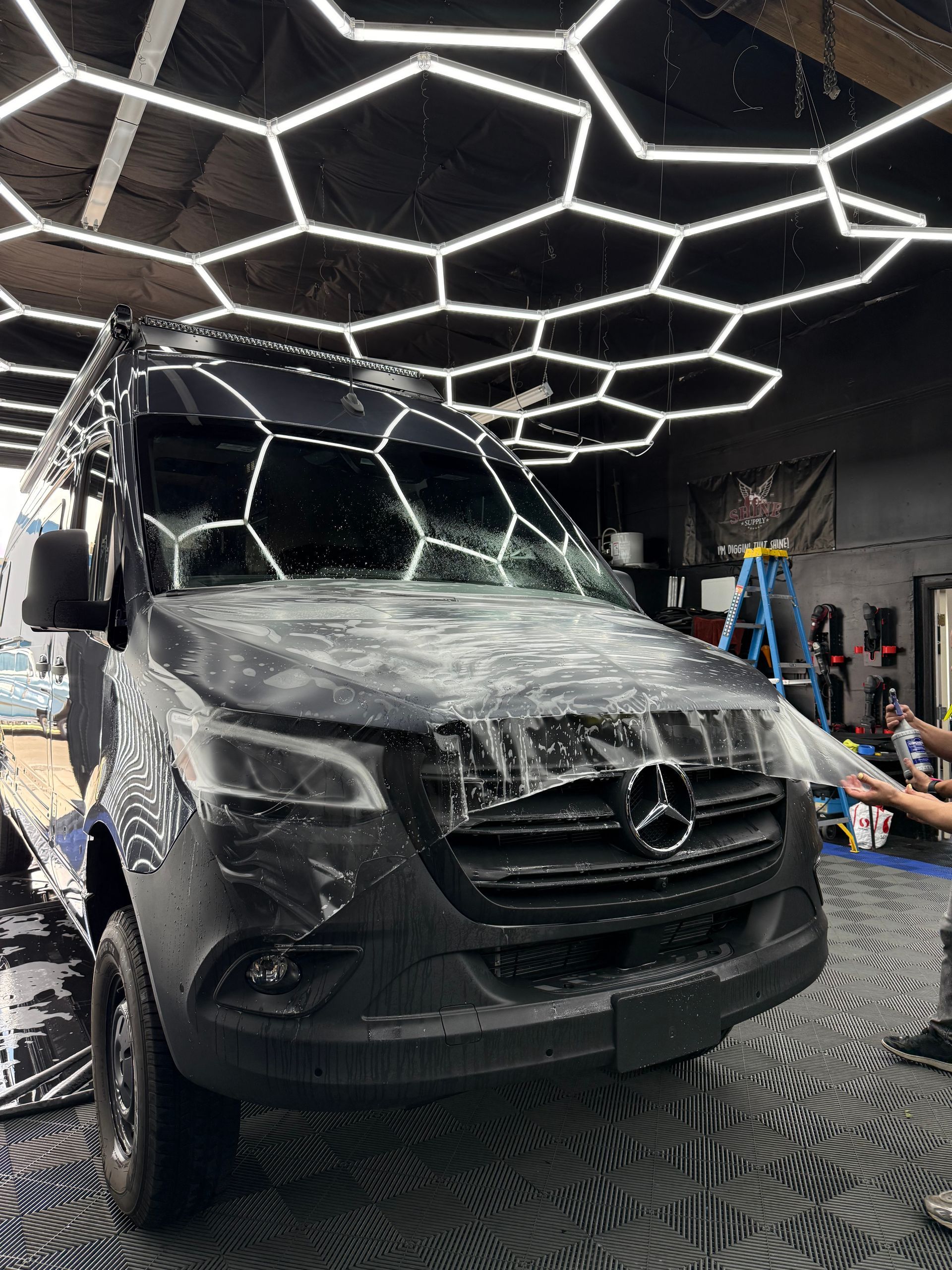 Black van covered in plastic wrap, under hexagon-shaped ceiling lights, inside a shop.