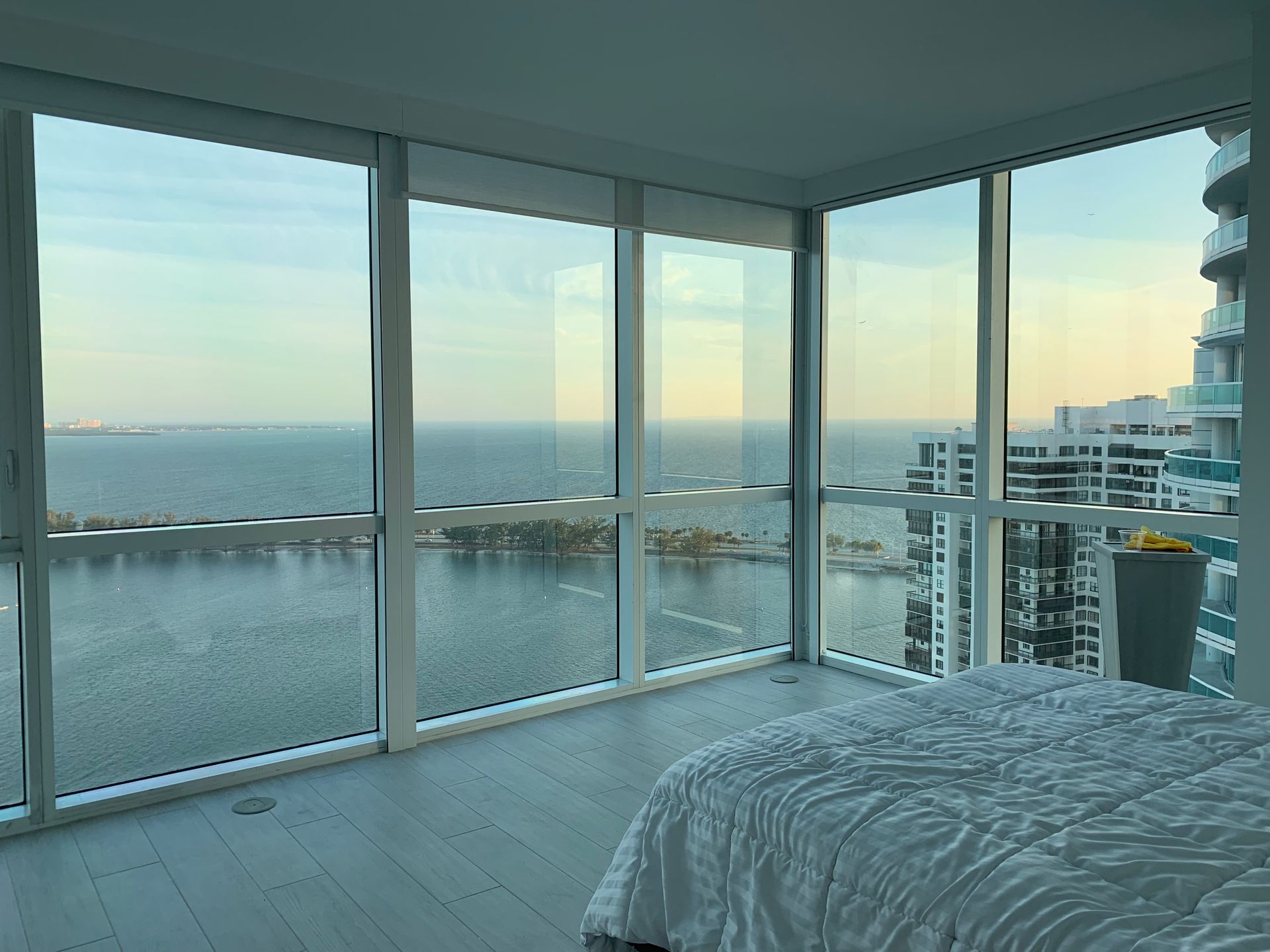 Bedroom with large windows overlooking a body of water and city buildings; bed in foreground.