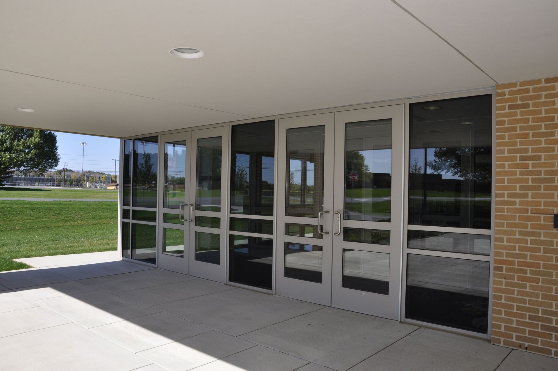 Glass doors under a covered walkway, leading to a building with a brick wall and green lawn visible.