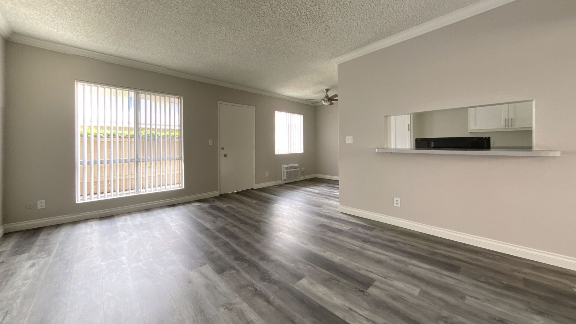 Empty, light-filled living room with gray wood-look flooring, neutral walls, and a pass-through to the kitchen.