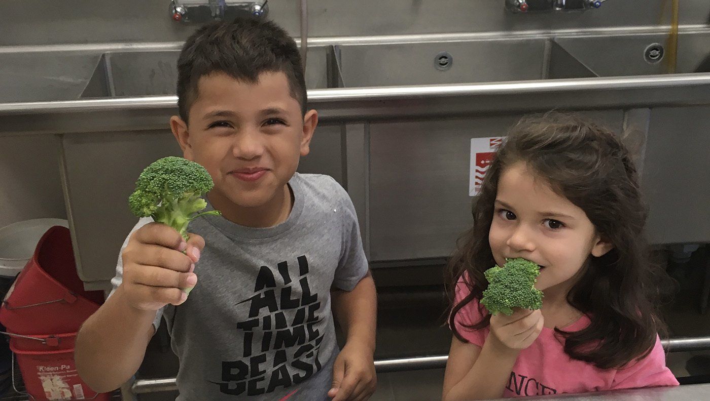 A boy and a girl are holding broccoli in front of a sink.