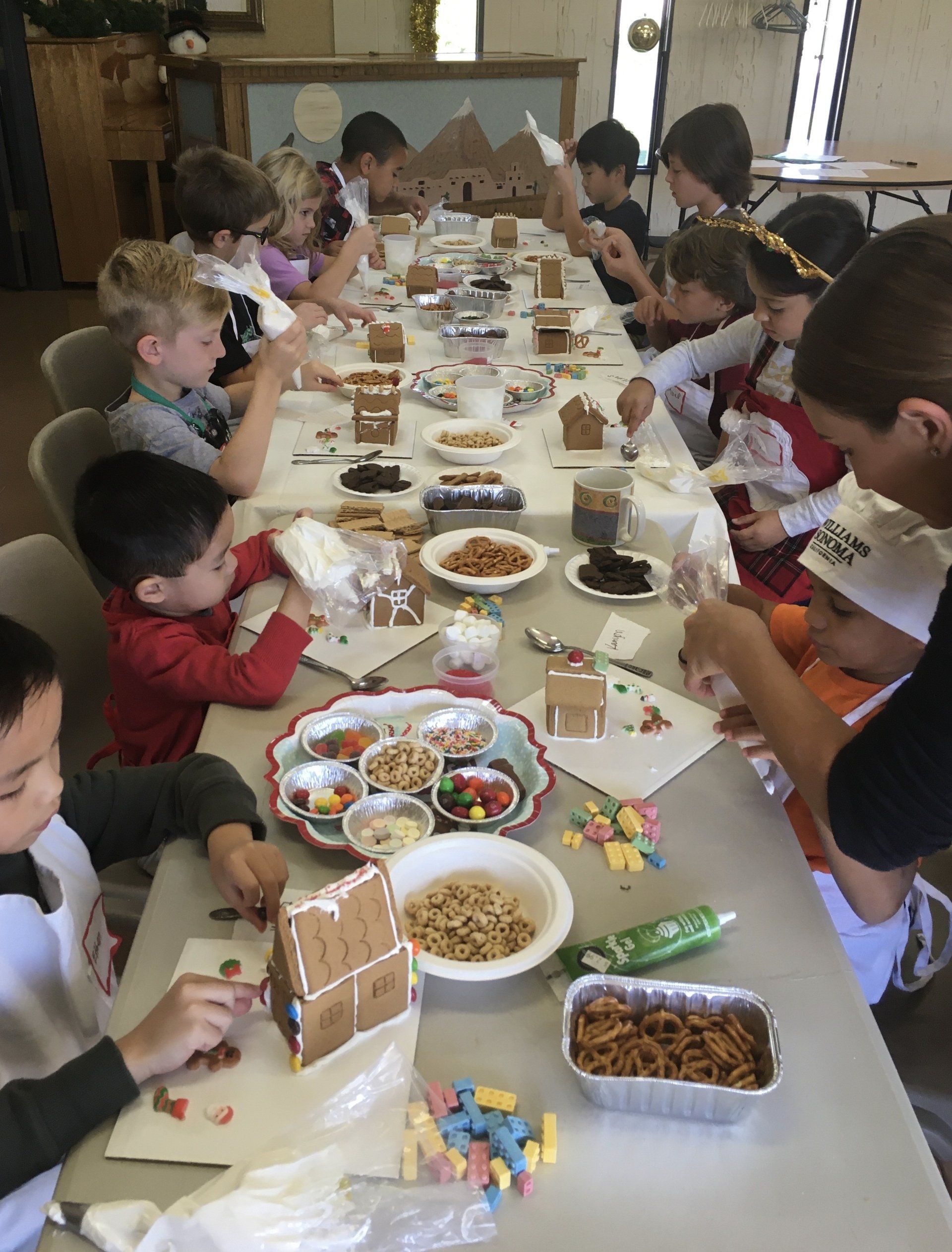 A group of children are sitting at a table decorating gingerbread houses.