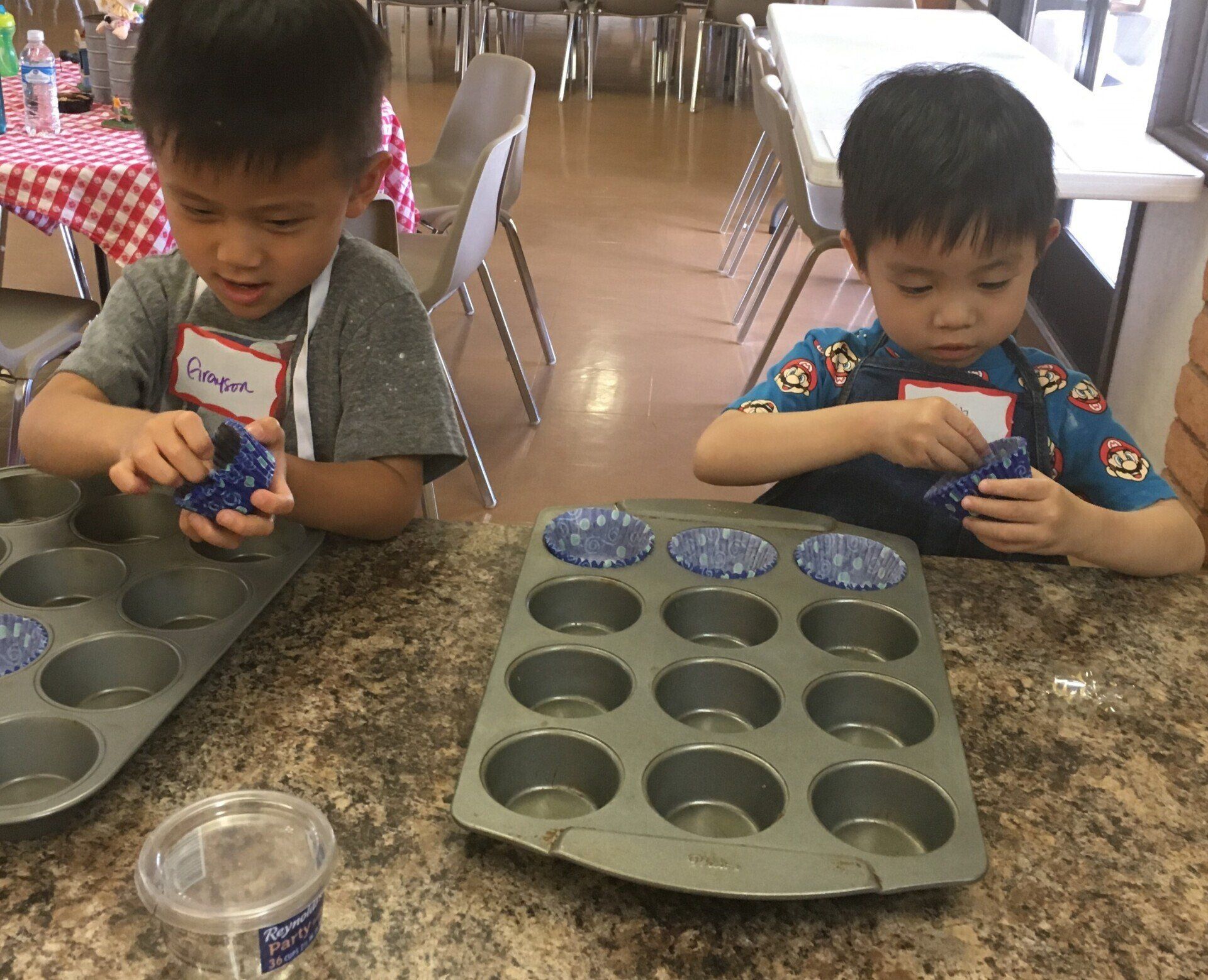 Two young boys are sitting at a table making cupcakes.