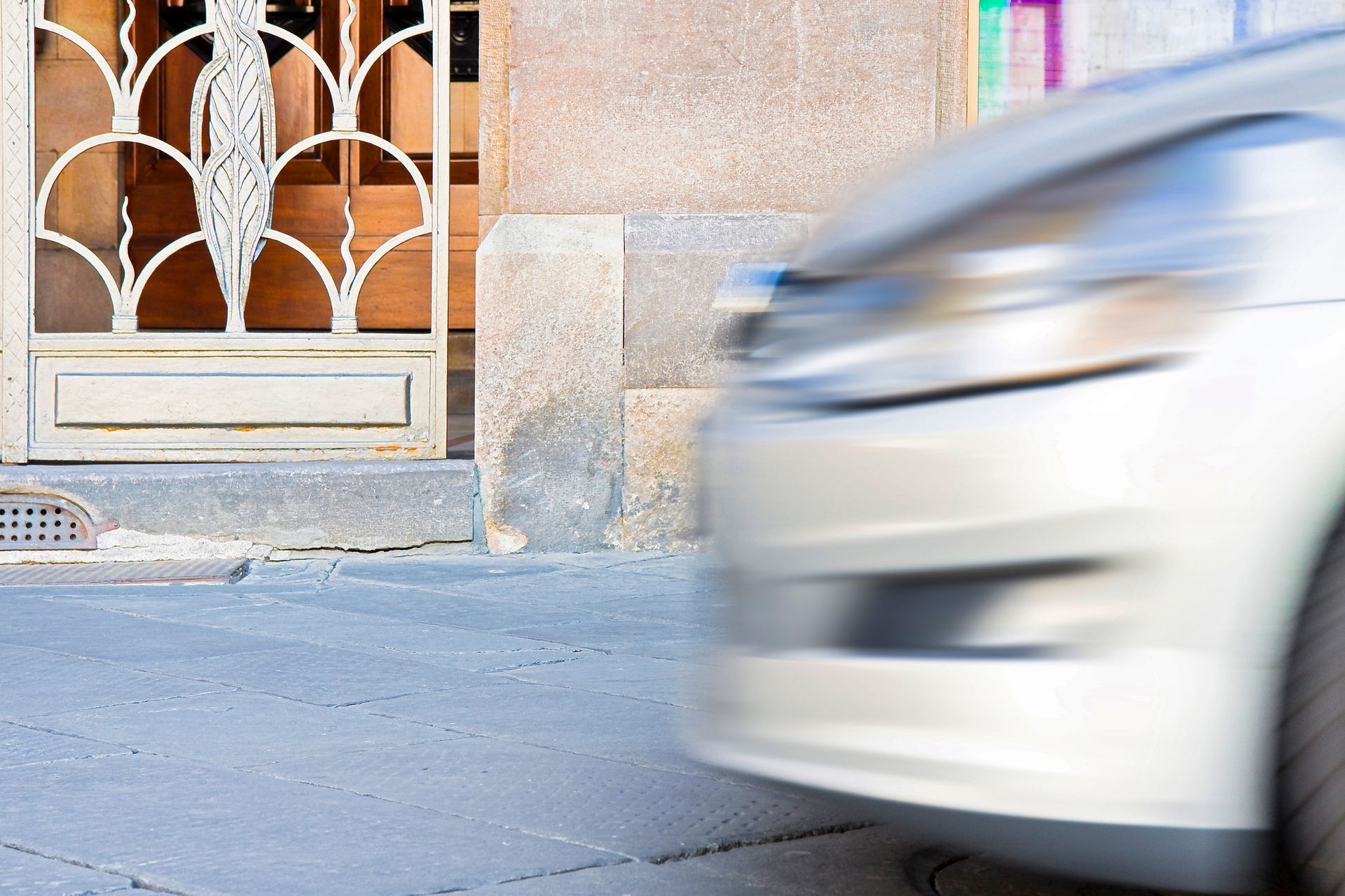 Un'auto argentata sfrecciava in movimento, sorpassando un edificio con un cancello metallico decorato.