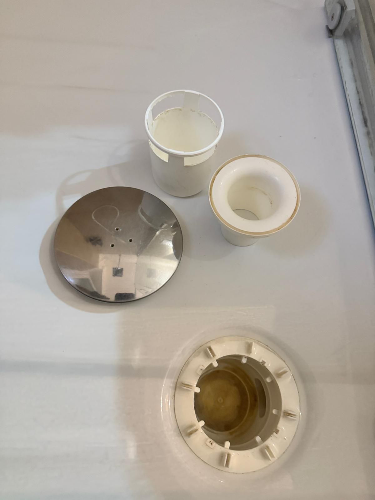 Componenti smontati dello scarico della doccia: cupola cromata, inserti bianchi e base su una superficie bianca.