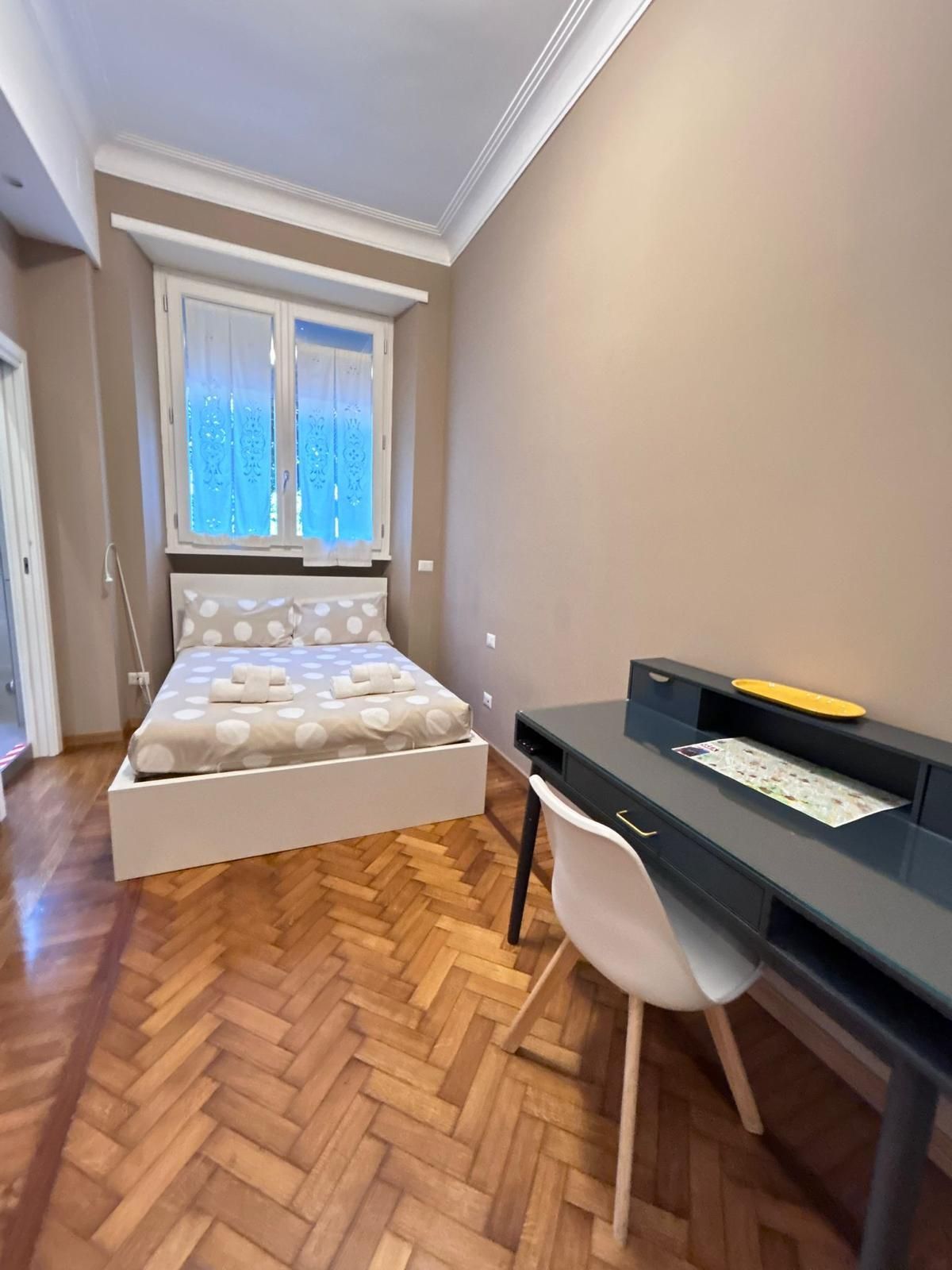 Camera da letto con letto, scrivania, sedia e finestra. Pareti color cuoio, pavimenti in legno chiaro e una sedia bianca.