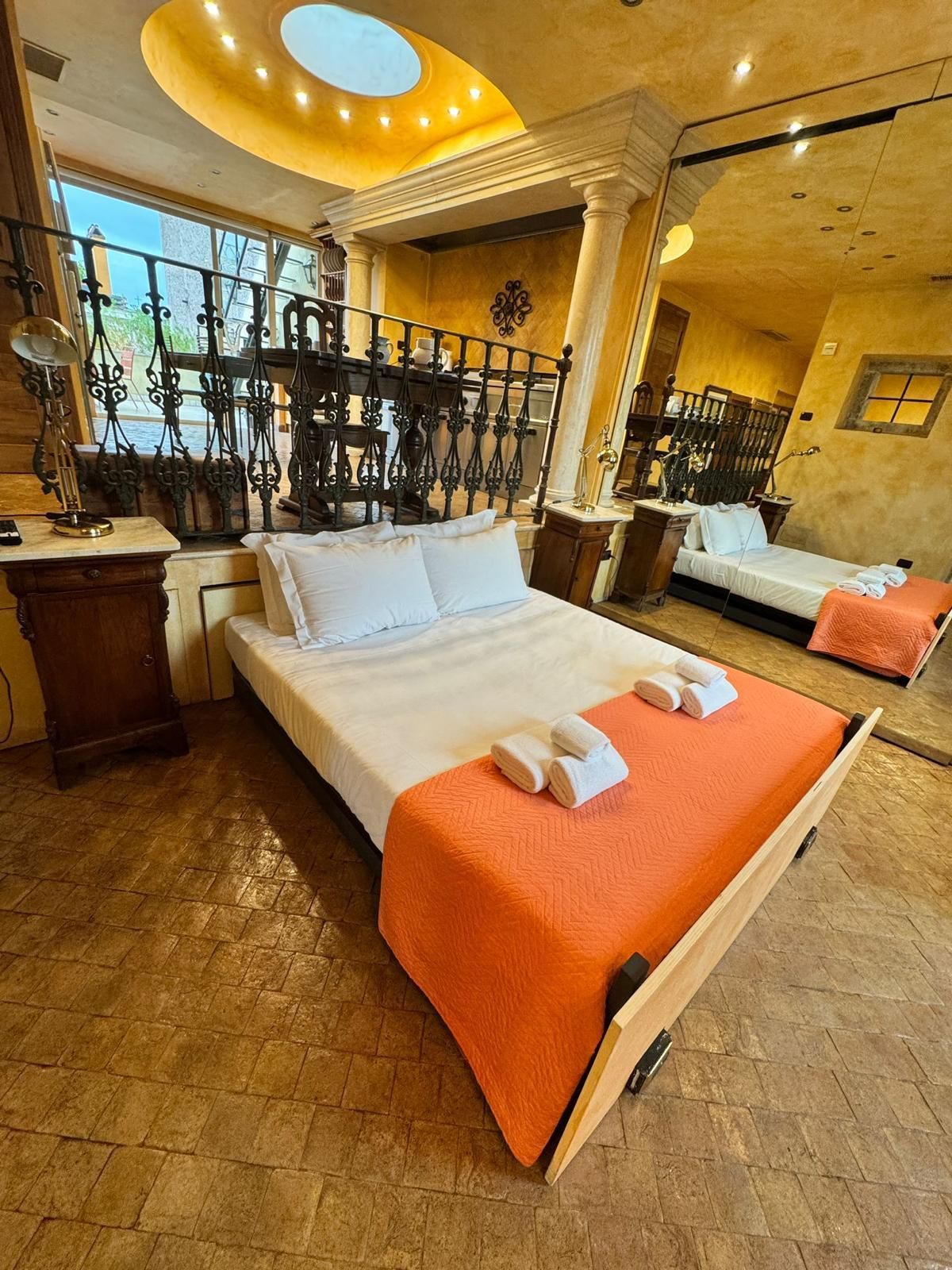 Camera da letto con letto, coperta arancione, cuscini bianchi e colonne, riflessi in uno specchio a parete.