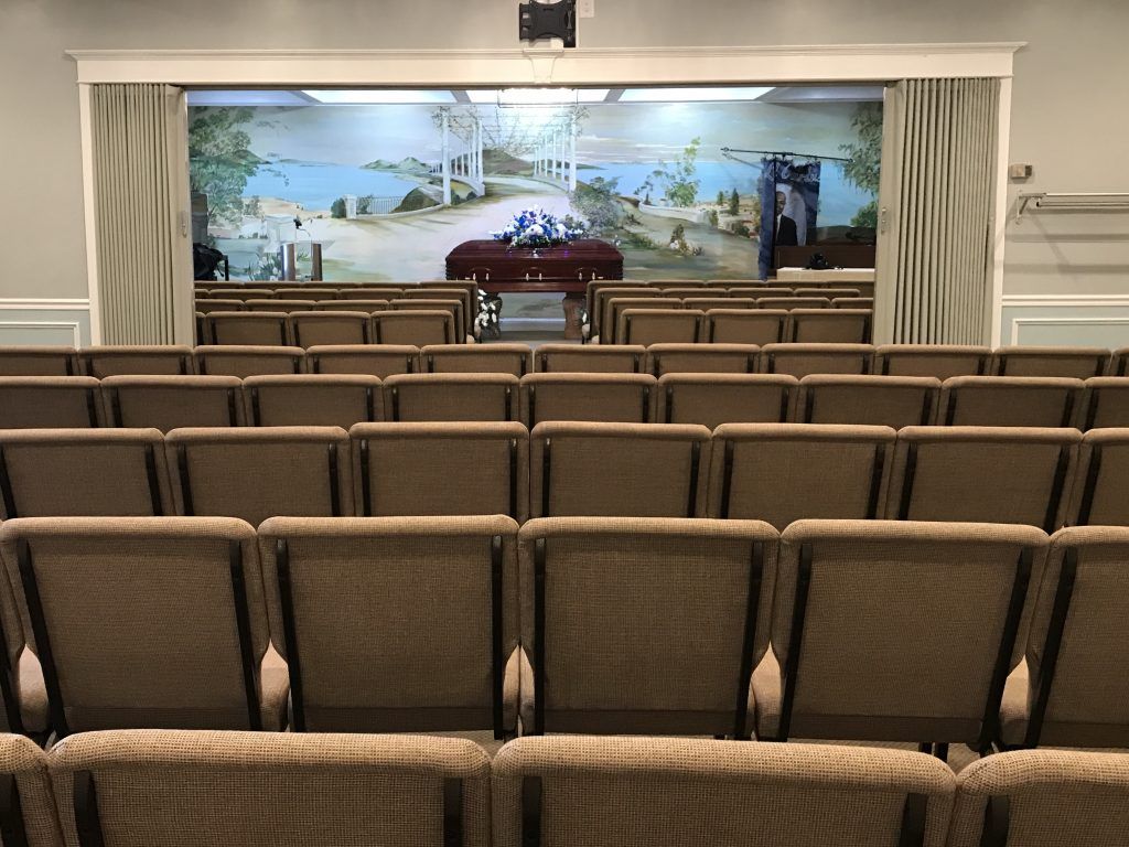 A well-lit room with rows of chairs, likely a funeral home chapel. Beige walls, carpeting, and ornate molding.