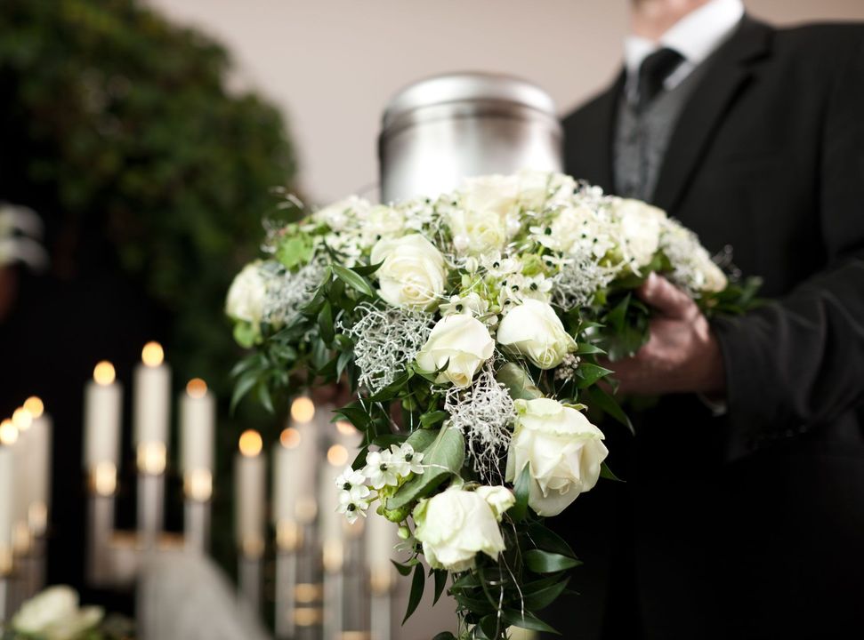 Man in a suit holding an urn with a floral arrangement at a funeral.  Candles and a green wreath are in the background.
