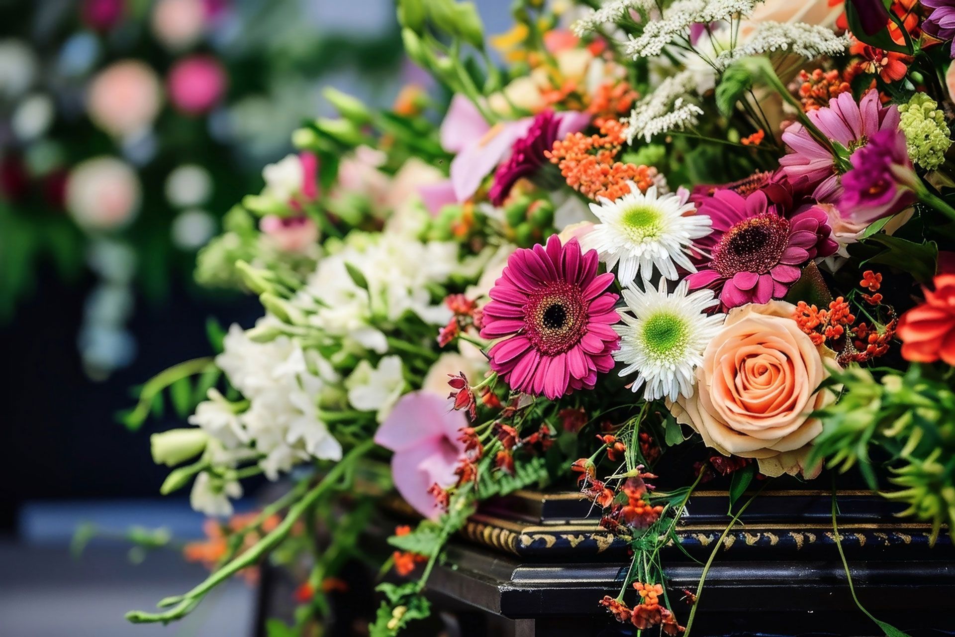 Colorful flower arrangement, including pink gerberas and orange roses, in front of a blurred background of more flowers.