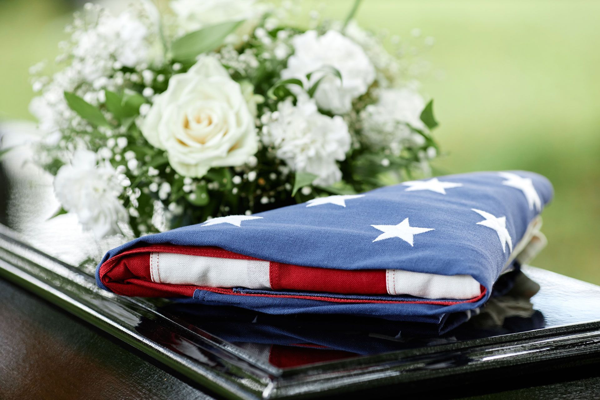 Folded American flag on a casket with a bouquet of white flowers in the background.