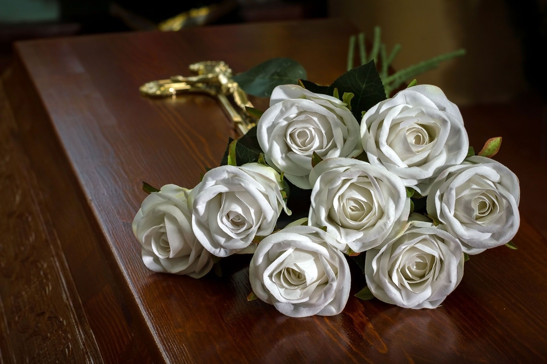 White roses and a gold crucifix rest on a closed, brown casket, likely at a funeral.
