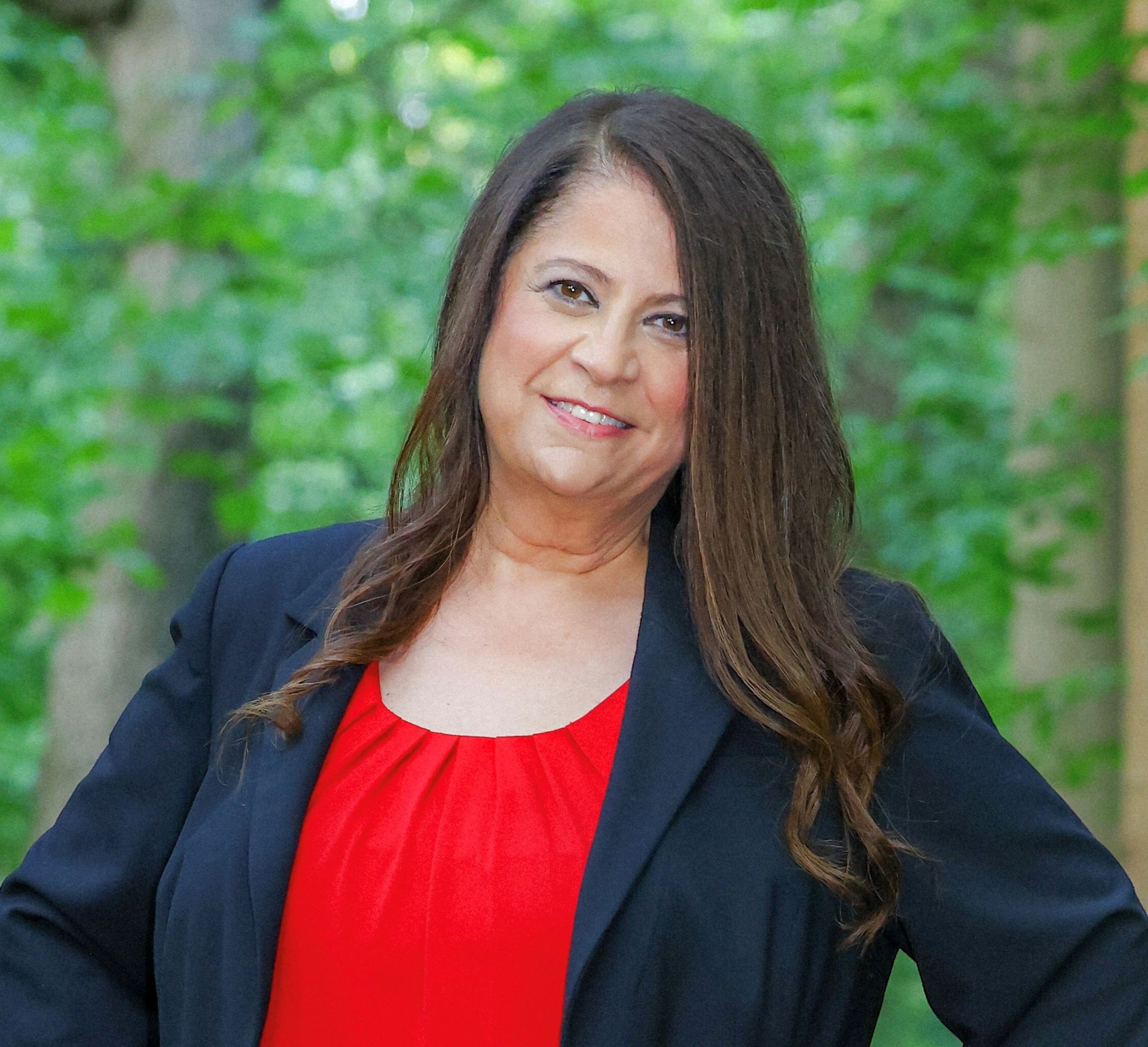 Pamela Price-Lerner wearing a red shirt and a black jacket is standing in front of trees .