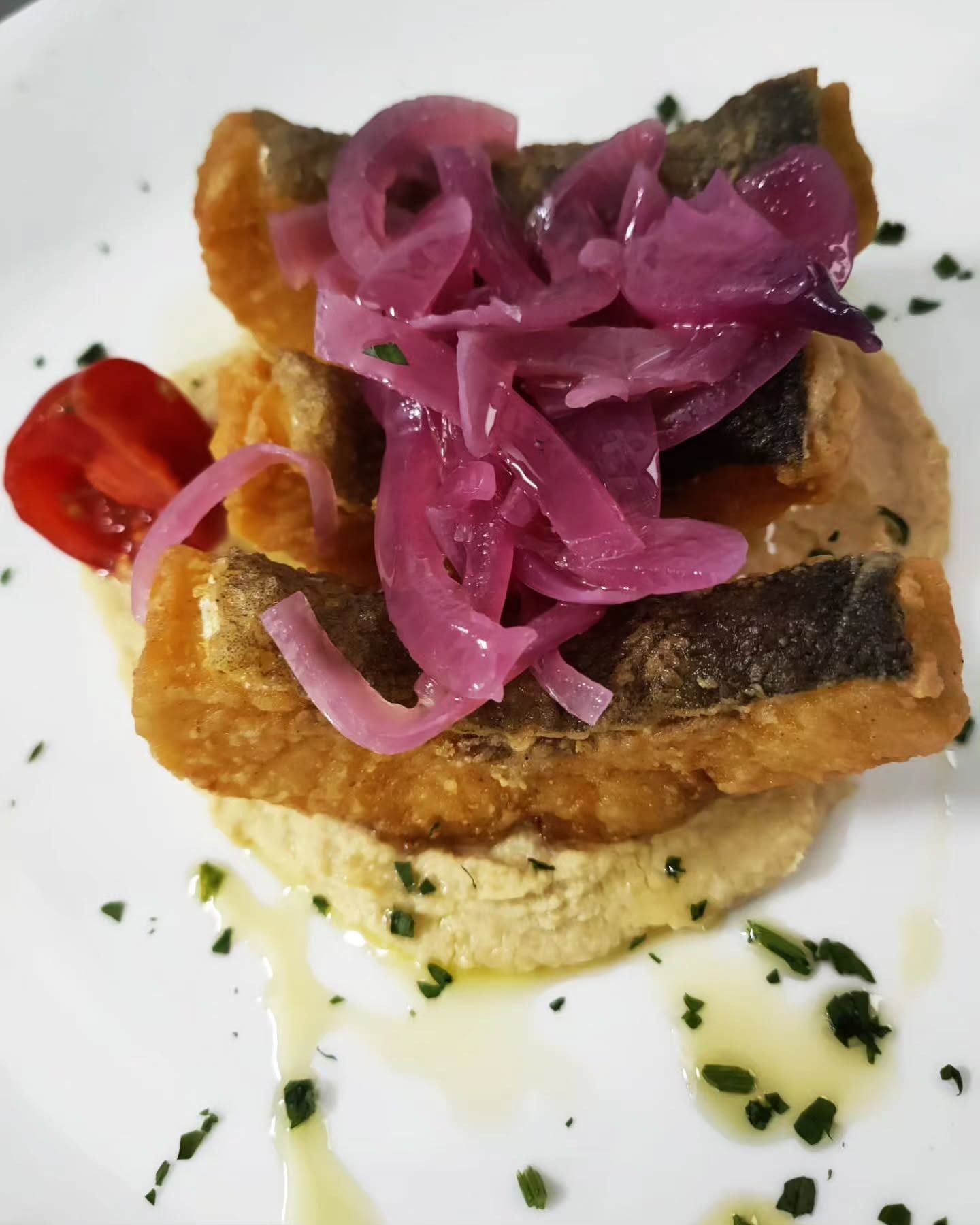Pesce fritto su hummus, condito con cipolle rosse sottaceto e un pomodoro, il tutto condito con un filo d'olio.