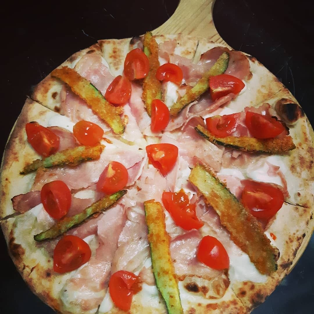Pizza con prosciutto, zucchine fritte e pomodorini su un tagliere di legno.