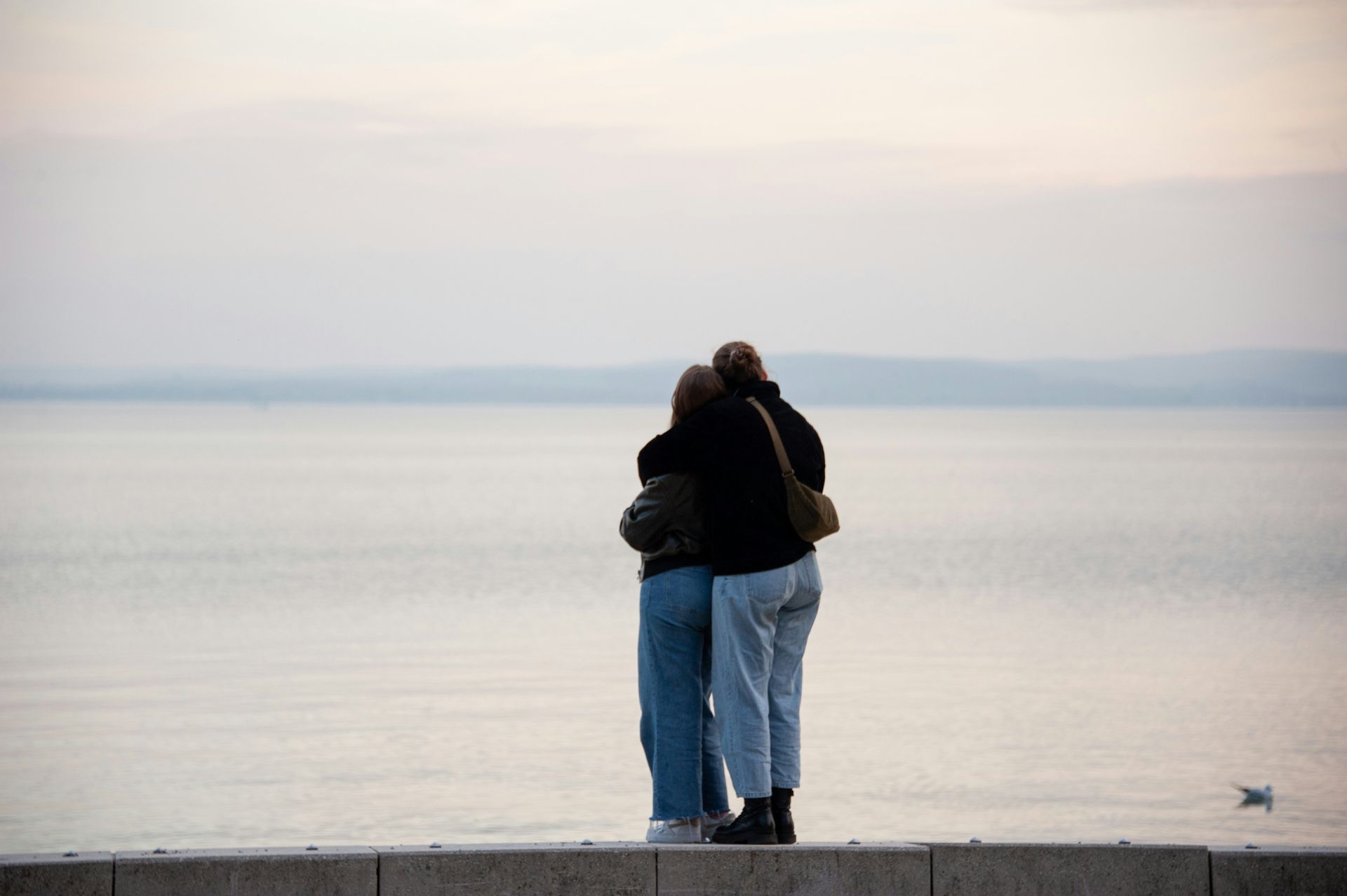 Two women are hugging each other while standing on a wall overlooking the ocean.