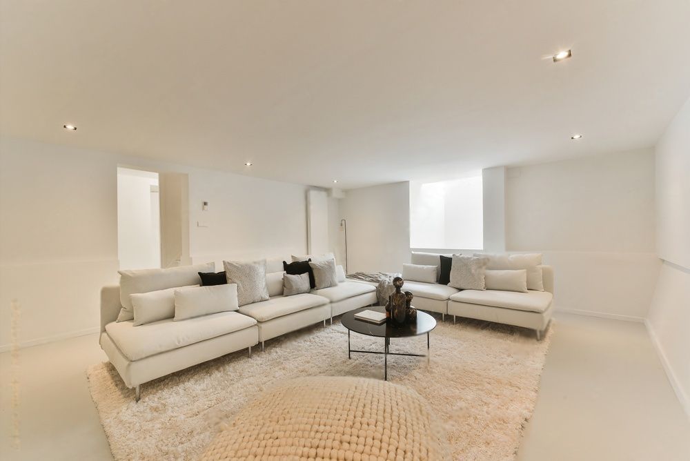 Een moderne, monochrome woonkamer met grote witte hoekbanken, een ronde salontafel en een zacht vloerkleed op een witte vloer.