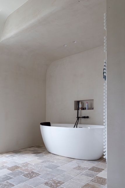 Een wit, vrijstaand bad tegen een minimalistische stucwand met een ingebouwde plank en tegelvloer in een moderne kamer.
