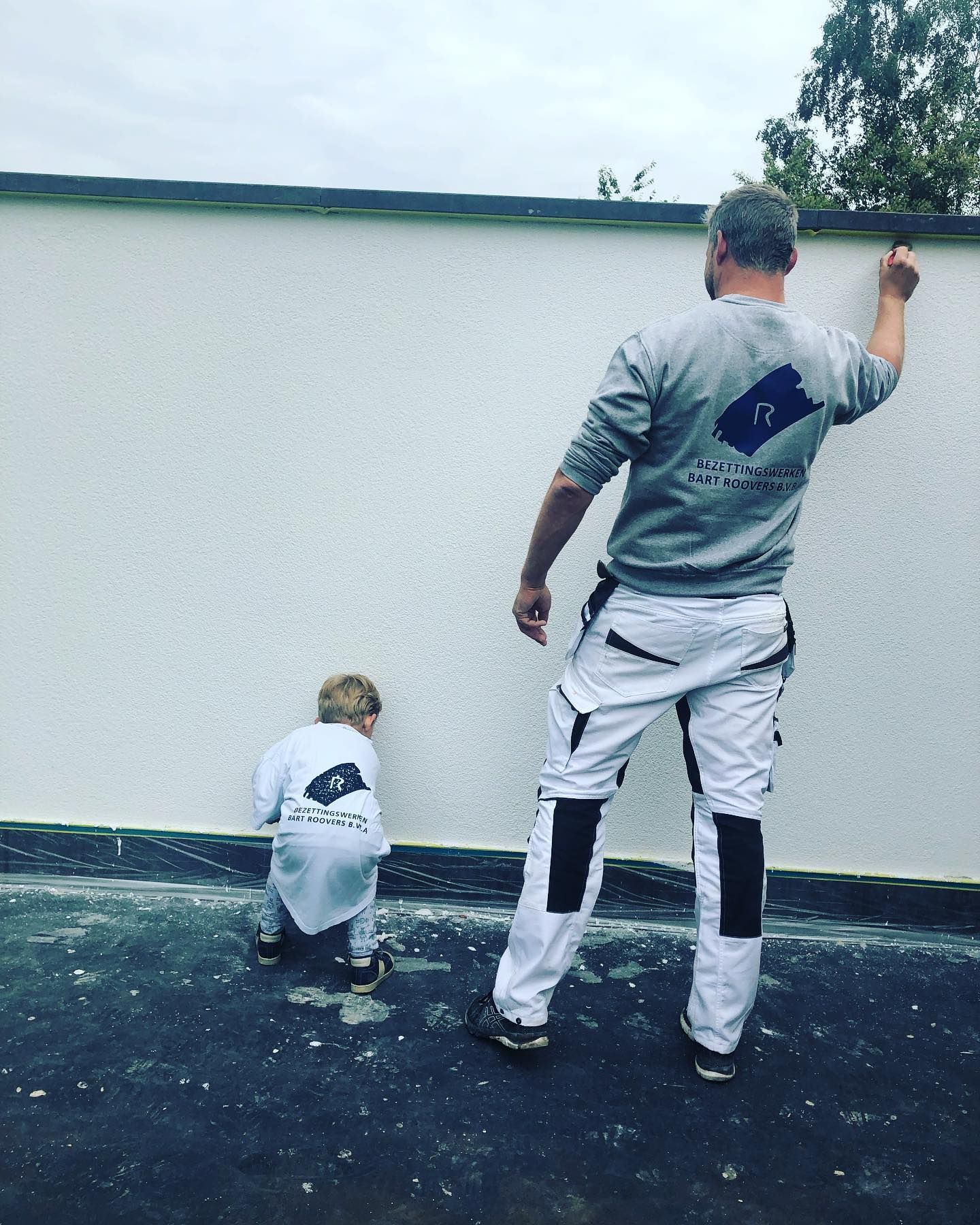 Een persoon en een kind, beiden gekleed in bijpassende grijze en witte werkkleding, schilderen een witte muur buiten op een donkere, gestructureerde ondergrond.