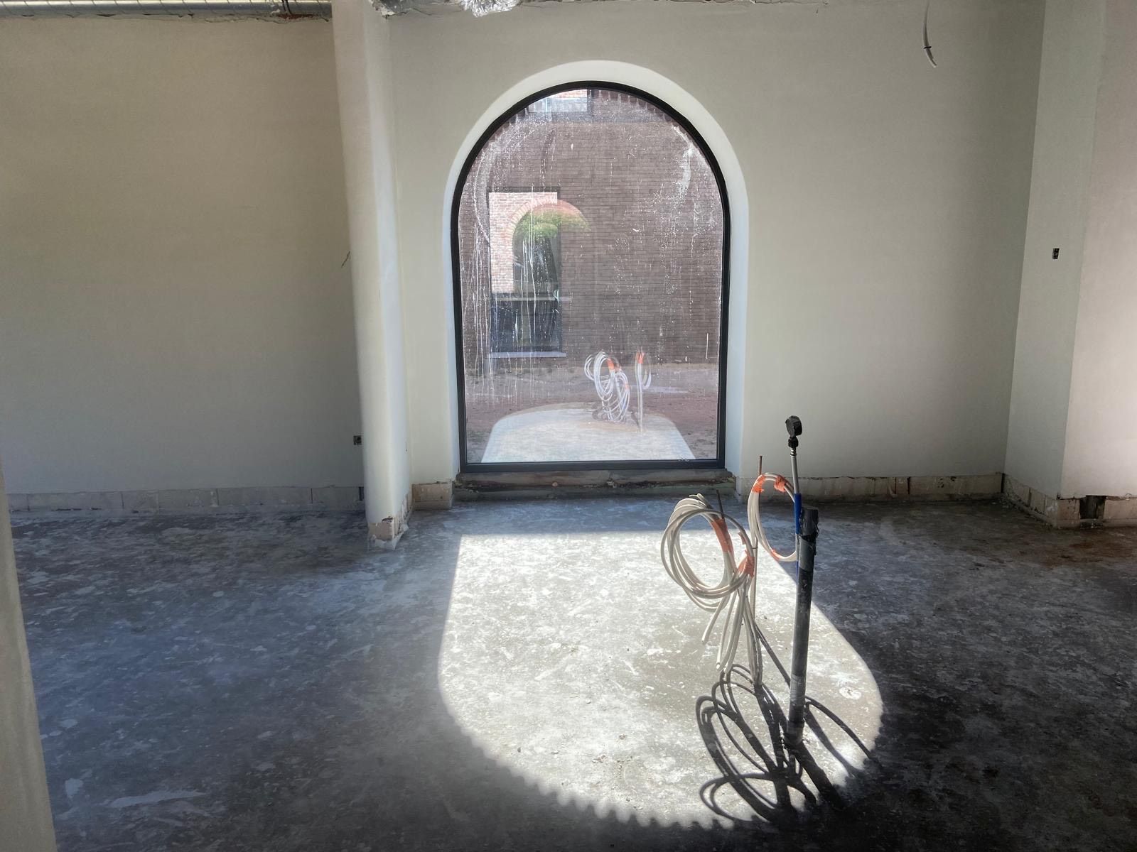 Een boogvormig raam laat licht binnen in een kamer met een onafgewerkte betonnen vloer en zichtbare elektrische bedrading.