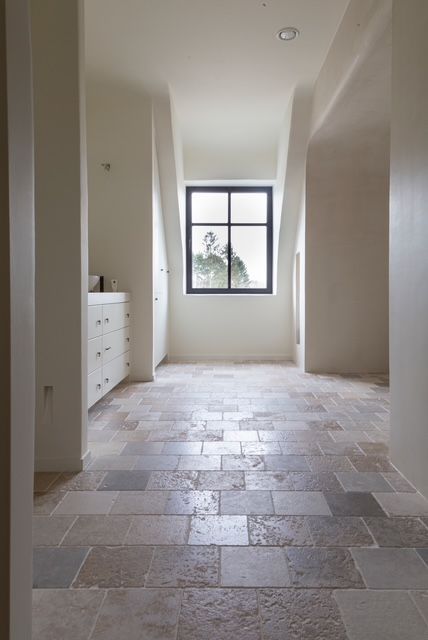 Een gang met een lichte stenen tegelvloer leidt naar een grote nis met ramen in een neutraal, minimalistisch interieur.