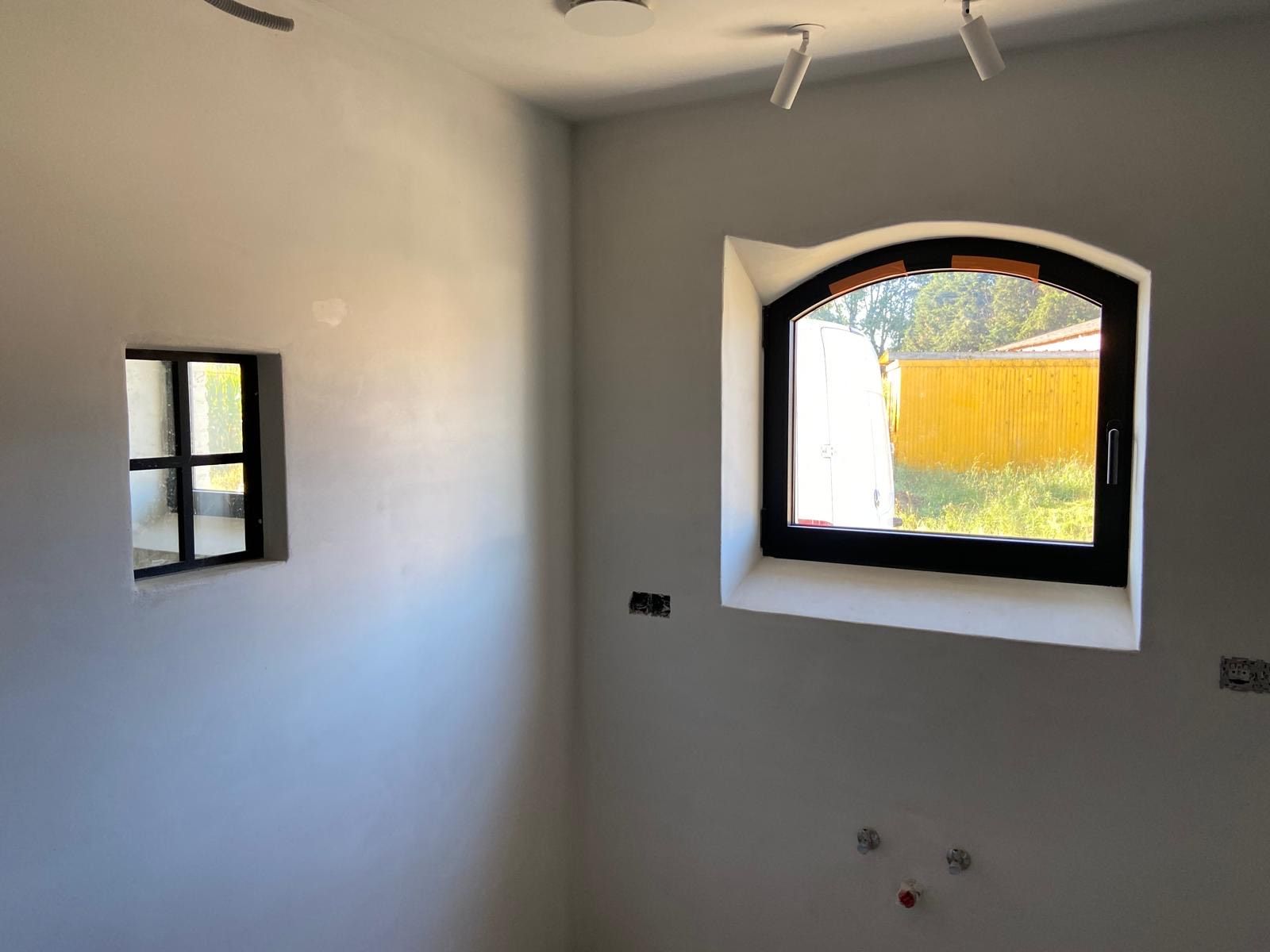 Een klein vierkant raam en een groot boogvormig raam met zwarte kozijnen in een witgeschilderde kamer met railverlichting aan het plafond.