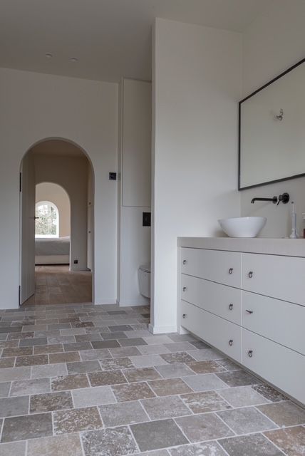 Een lichte, minimalistische badkamer met stenen tegelvloer, een wit badmeubel, een ronde wasbak en gewelfde deuropeningen aan de achterzijde.
