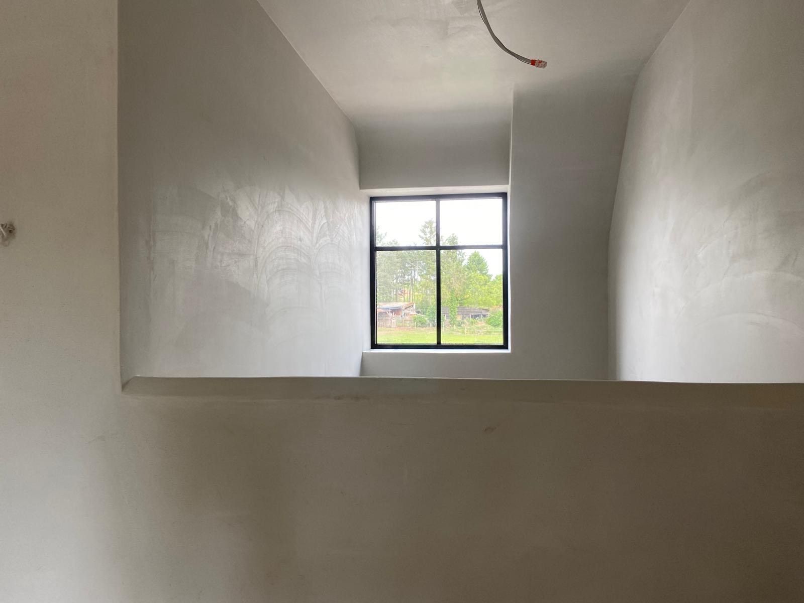 Een interieurfoto van een kamer met witte muren, een grote opening die uitzicht biedt op een andere ruimte, en een raam met uitzicht op groen.