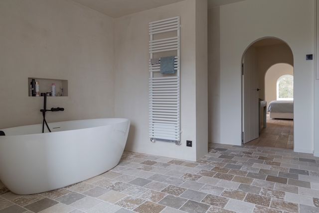 Een moderne badkamer met een vrijstaand wit bad, tegelvloer, witte wandradiator en een boogvormige deuropening naar een slaapkamer.