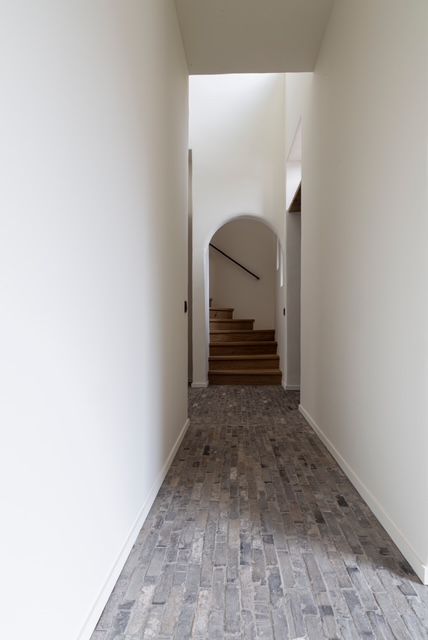 Een gang met witte muren en een grijze, gestructureerde bakstenen vloer, die via een boogvormige deuropening toegang geeft tot een houten trap.