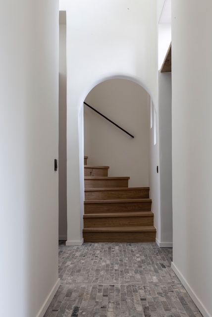 Een gang leidt via een witte boog naar een houten trap. De vloer is bedekt met verweerde grijze bakstenen.