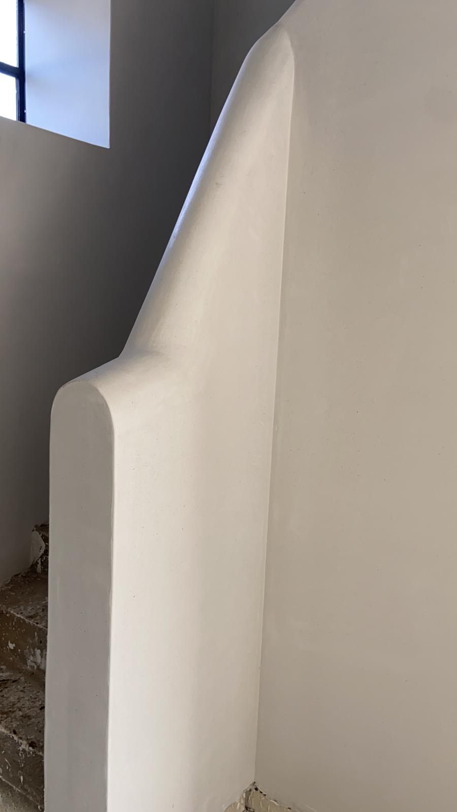 Een gladde, witte, afgeronde gipsen muurconstructie, mogelijk een trapleuning, tegen een effen witte binnenmuur.