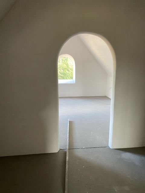 Een boogvormige deuropening leidt naar een lichte, onafgewerkte zolderkamer met een rond raam en een lichtgrijze vloer.