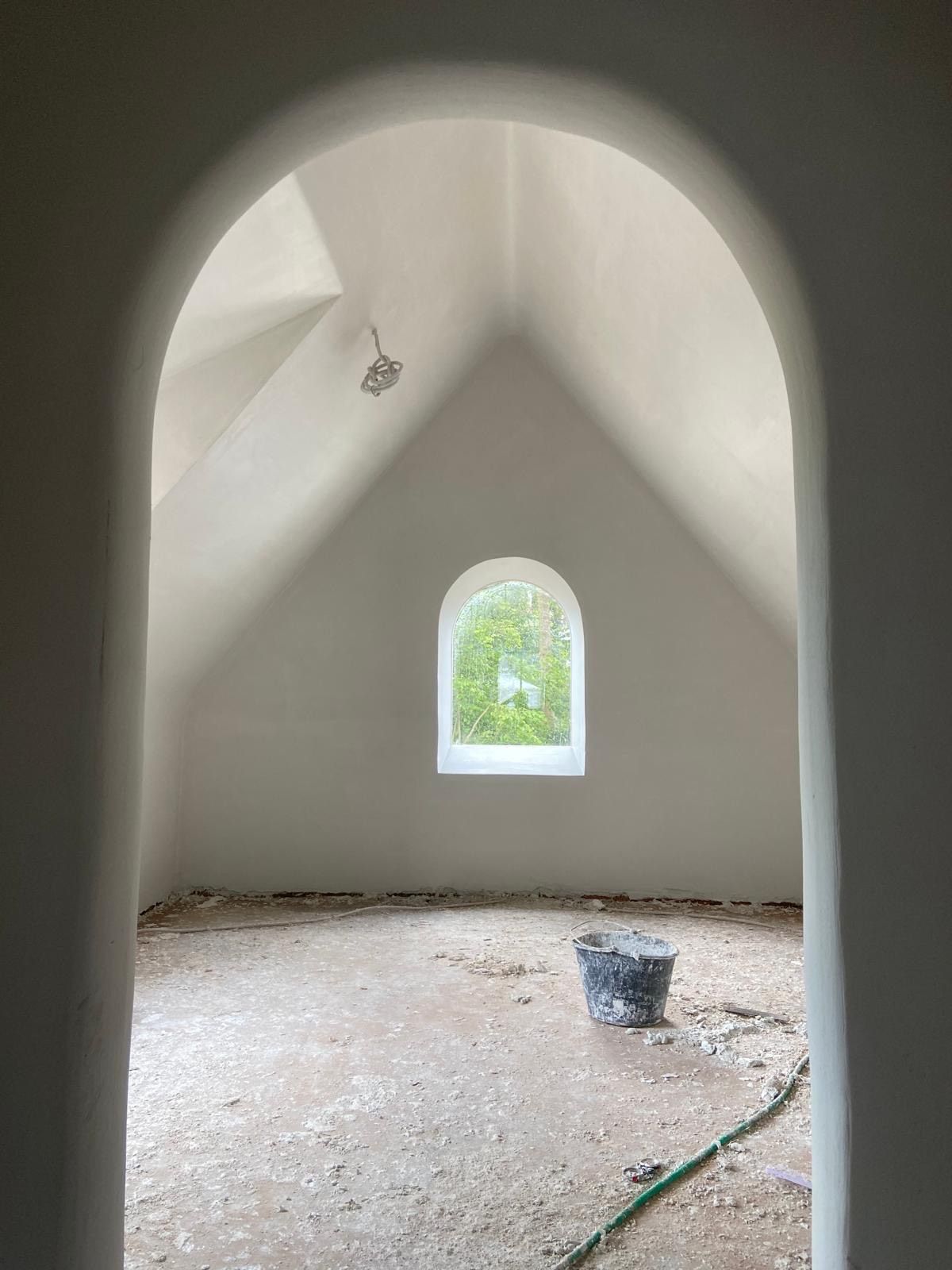 Een onafgewerkte kamer, zichtbaar door een grote boog, met witgepleisterde muren, een puntig plafond en een boogvormig raam.
