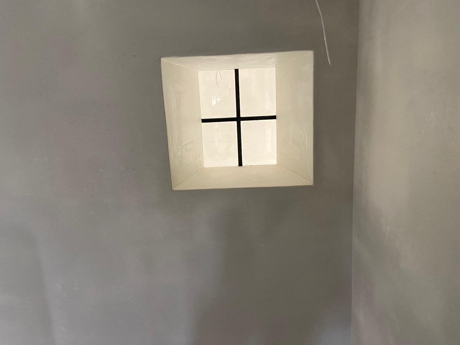 Een klein, vierkant raam met een wit frame en een zwarte kruisvormige raamstijl, gecentreerd in een effen grijze muur.