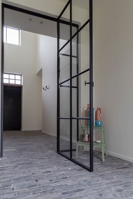 In een hal met grijze tegelvloer staat een grote glazen deur met zwart frame open, naast een klein plankje met vazen.