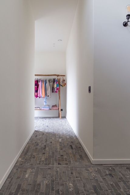 Een witte gang met een grijze stenen vloer leidt naar een houten rek waaraan kleurrijke kleding hangt.