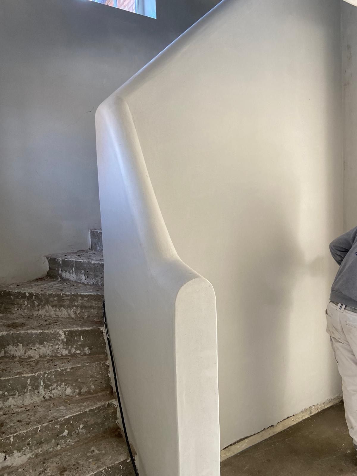 Een gladde, witgepleisterde, gebogen muur scheidt een onafgewerkte betonnen trap van een persoon die aan de rechterkant staat.