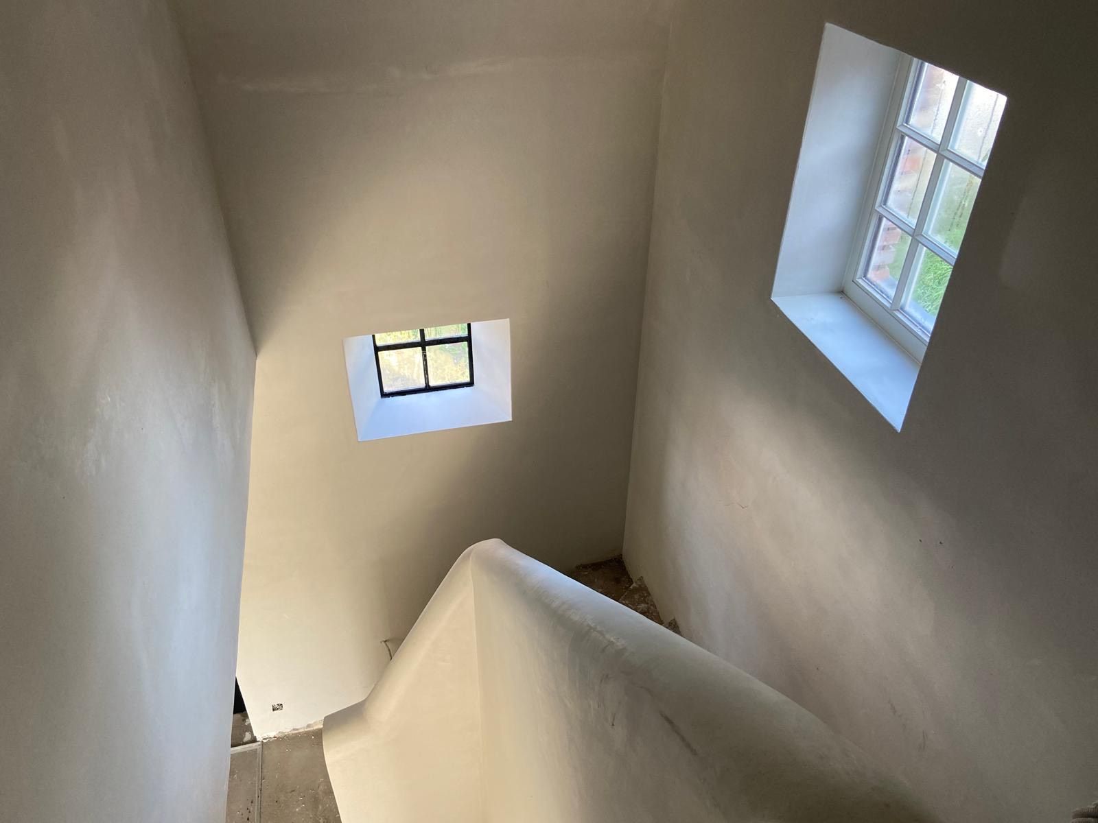 Uitzicht op een trappenhuis met gladde, witgepleisterde muren en twee kleine ramen met roedeverdeling op verschillende hoogtes.