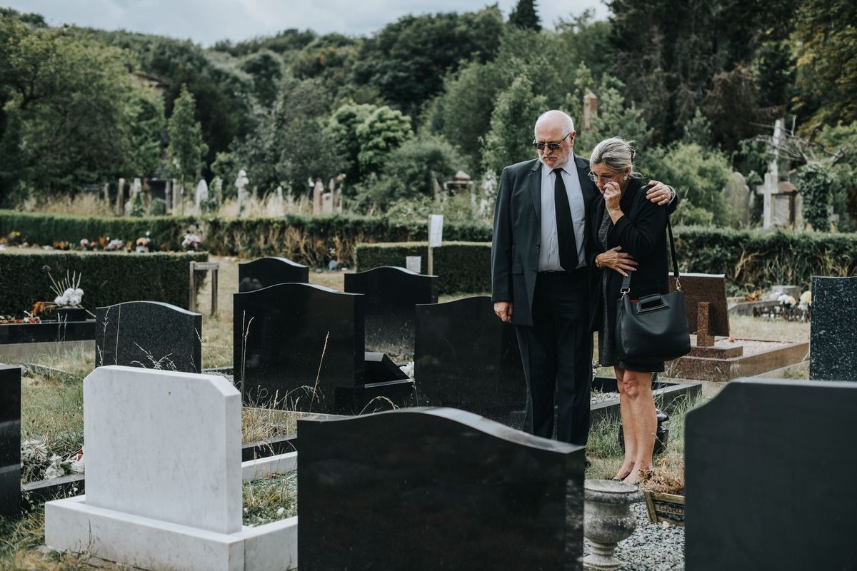 Man comforts woman at a cemetery, graves in view, overcast sky.