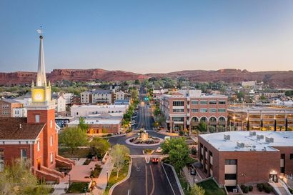 Aerial view of a city street lined with buildings and a church with a tall steeple, set against red rock mountains.