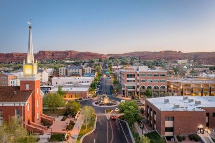 Aerial view of a city street lined with buildings and a church with a tall steeple, set against red rock mountains.