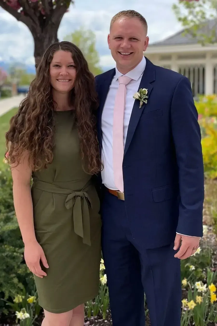 Woman in olive dress and man in navy suit, standing together. Both are smiling outdoors.