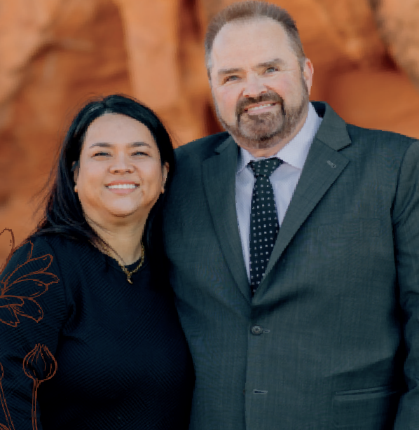 Woman in olive dress and man in navy suit, standing together. Both are smiling outdoors.