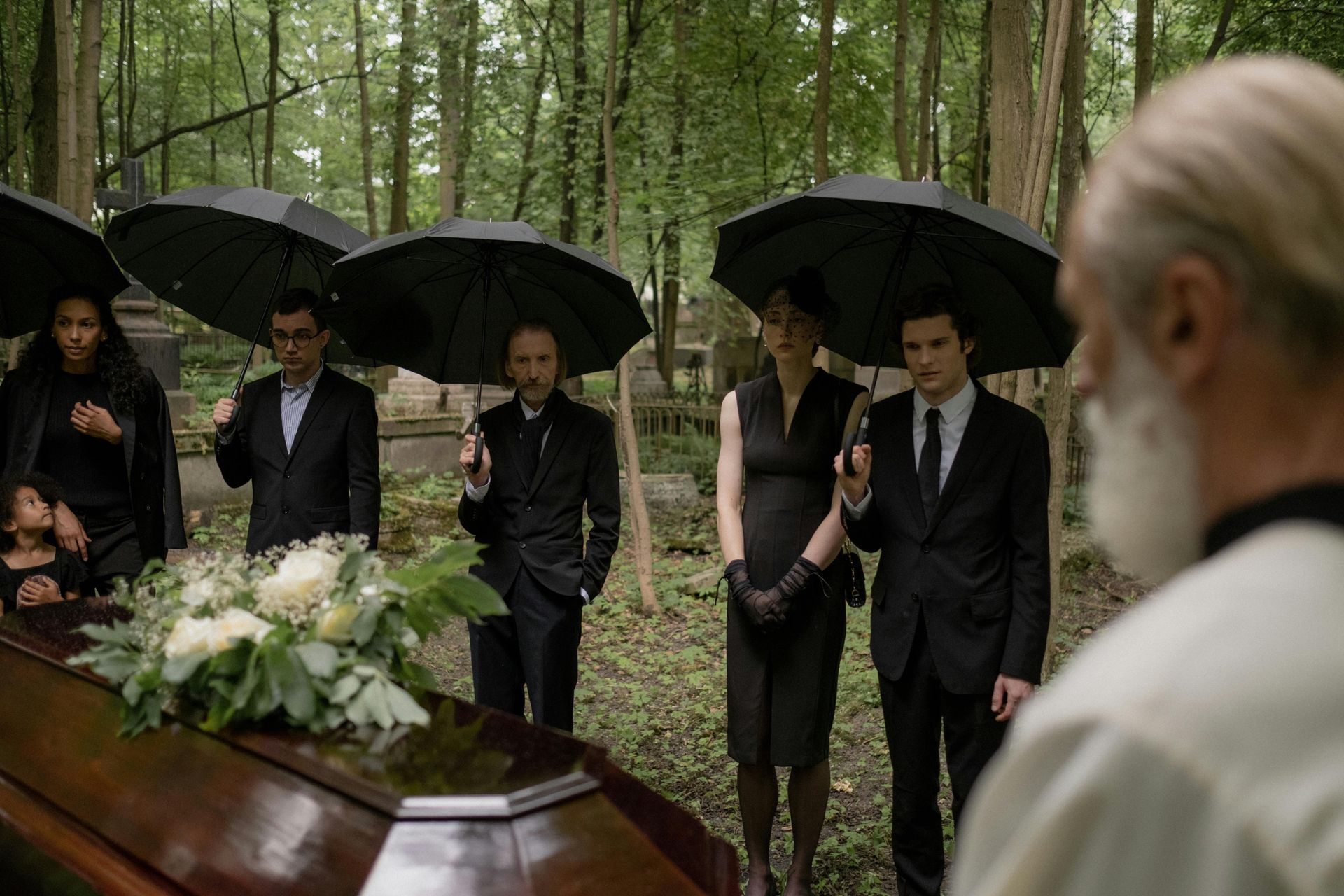 People with umbrellas stand at a funeral, coffin in front. Trees and foliage surround them.