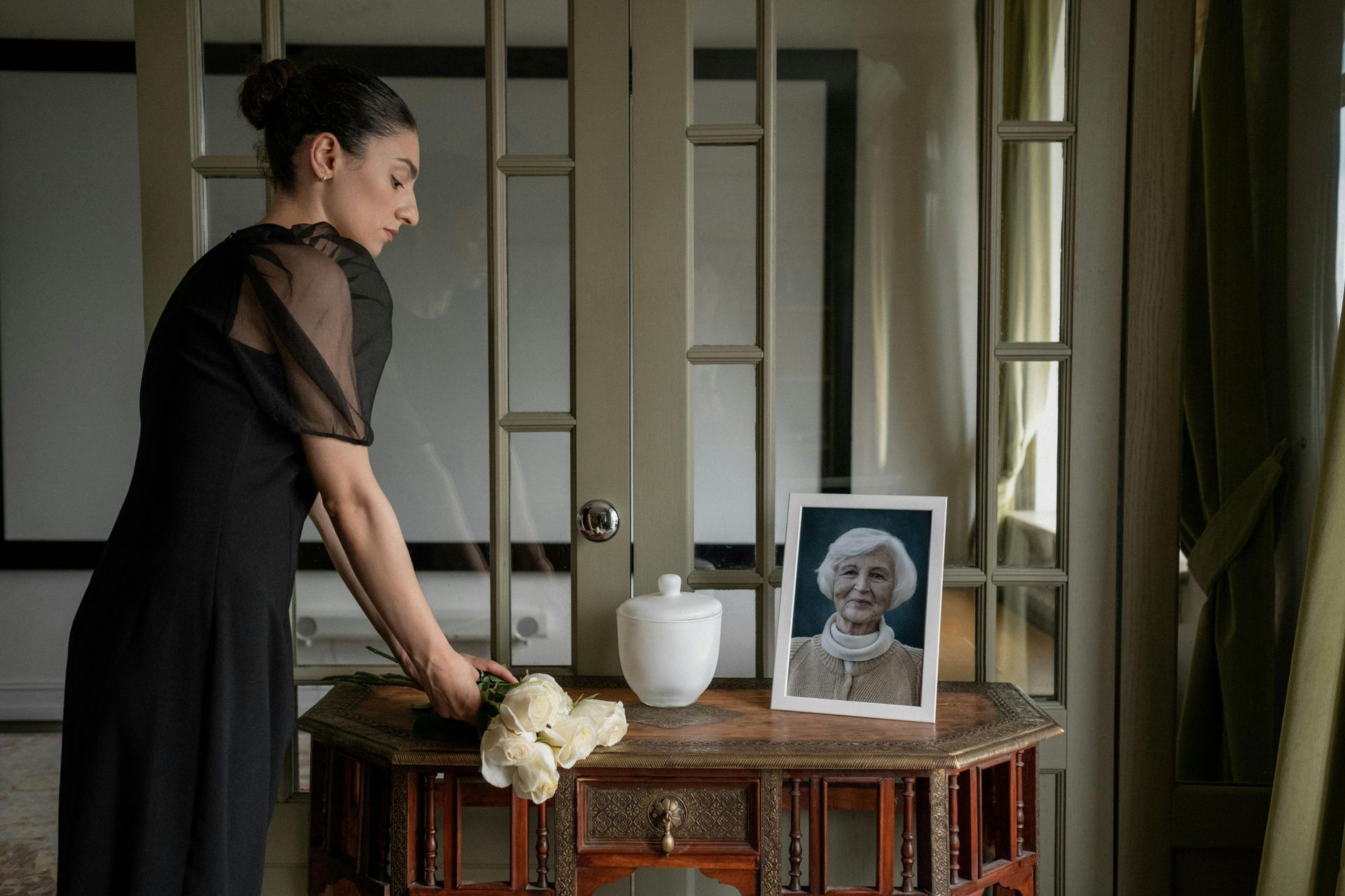 Woman in black dress arranging flowers near a portrait and urn, indoors.