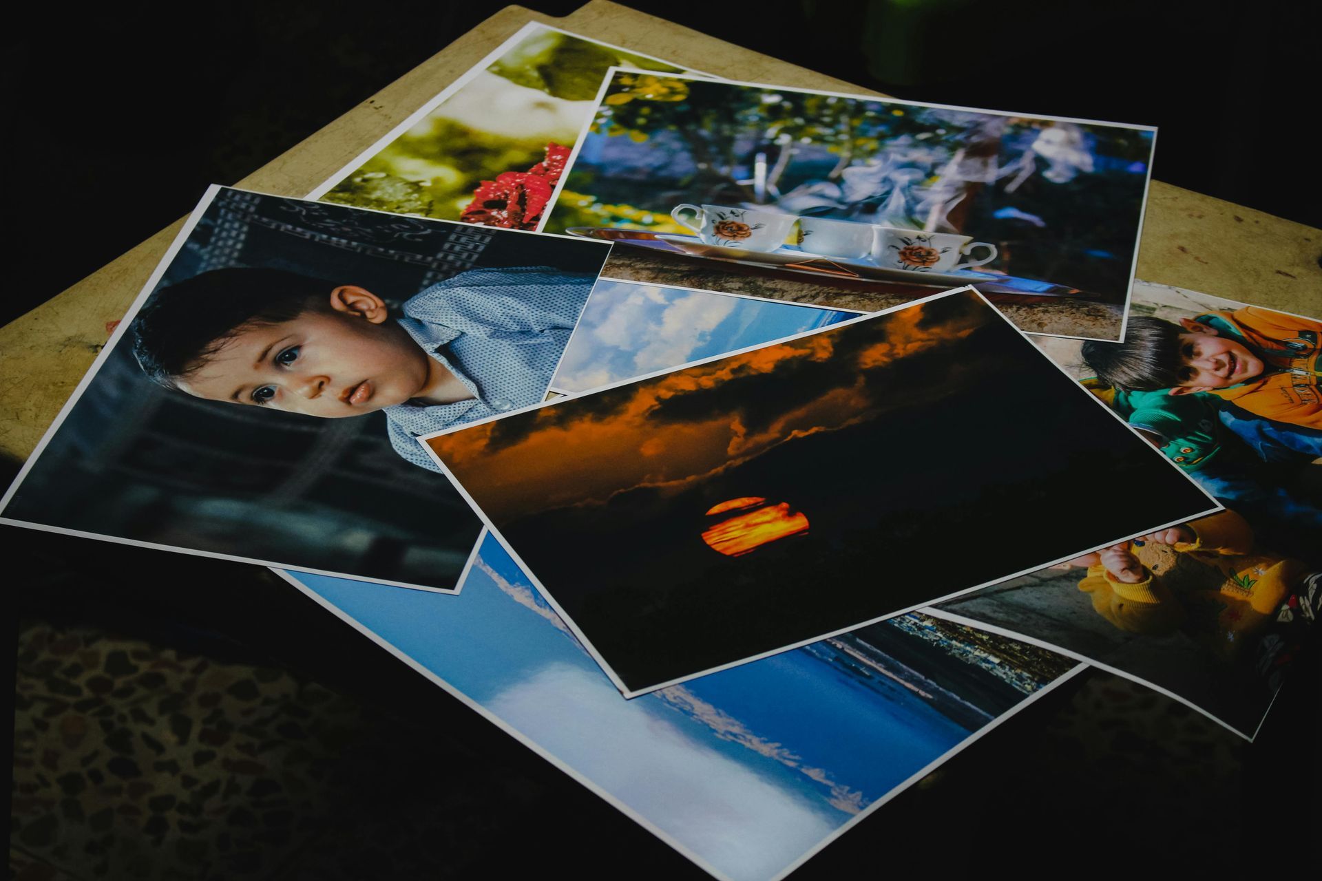 Pile of photos: child, sunset, tea set, outdoor scenes, and water reflections on a table.