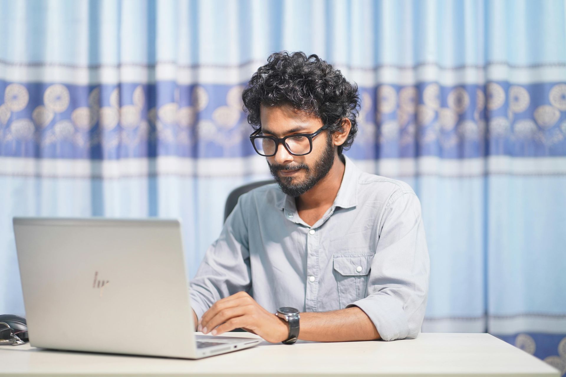 Man with glasses, shirt, and watch, working on a laptop at a desk in front of a blue curtain.