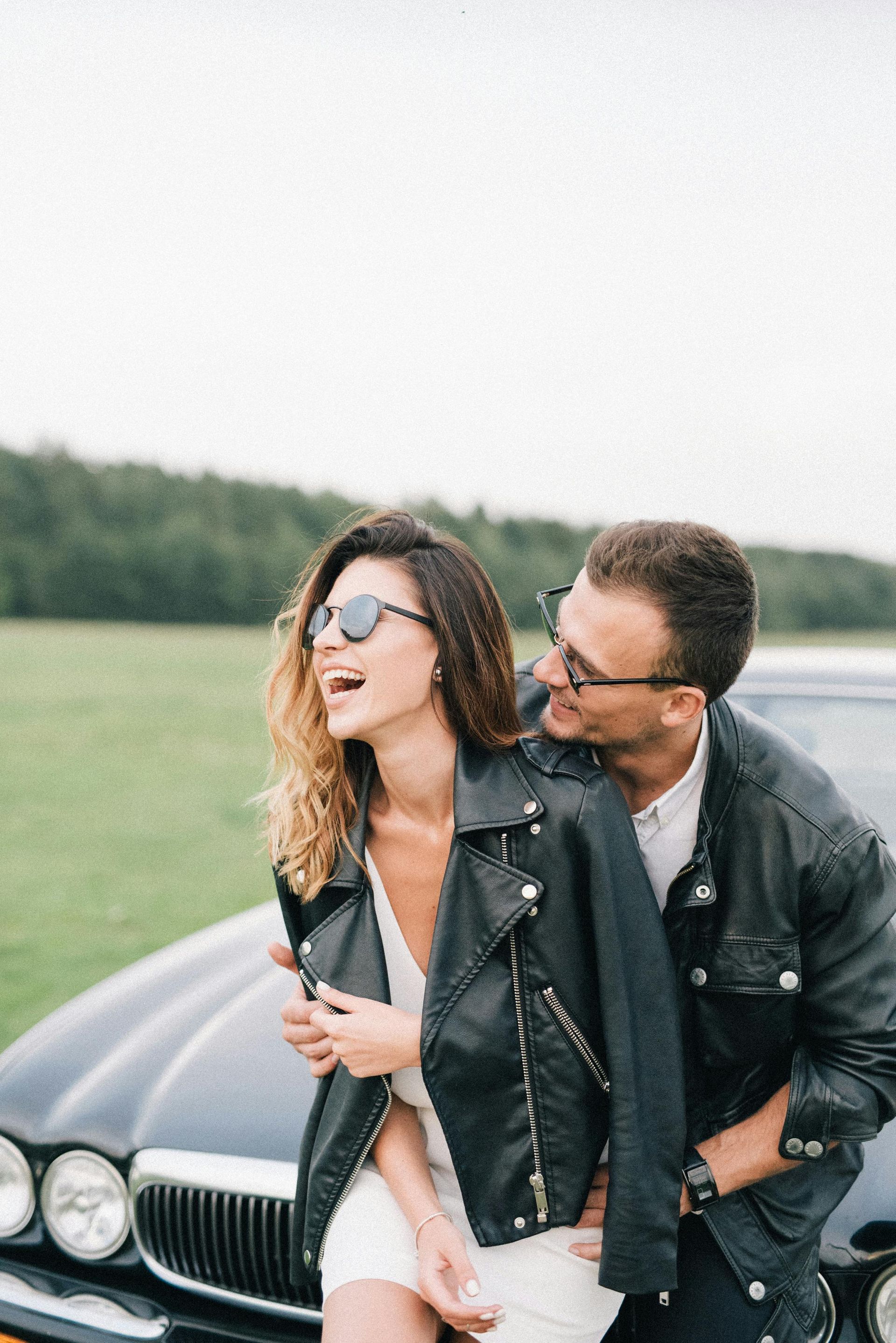 Couple laughing, embracing by a black car in a field; both wearing sunglasses and leather jackets.
